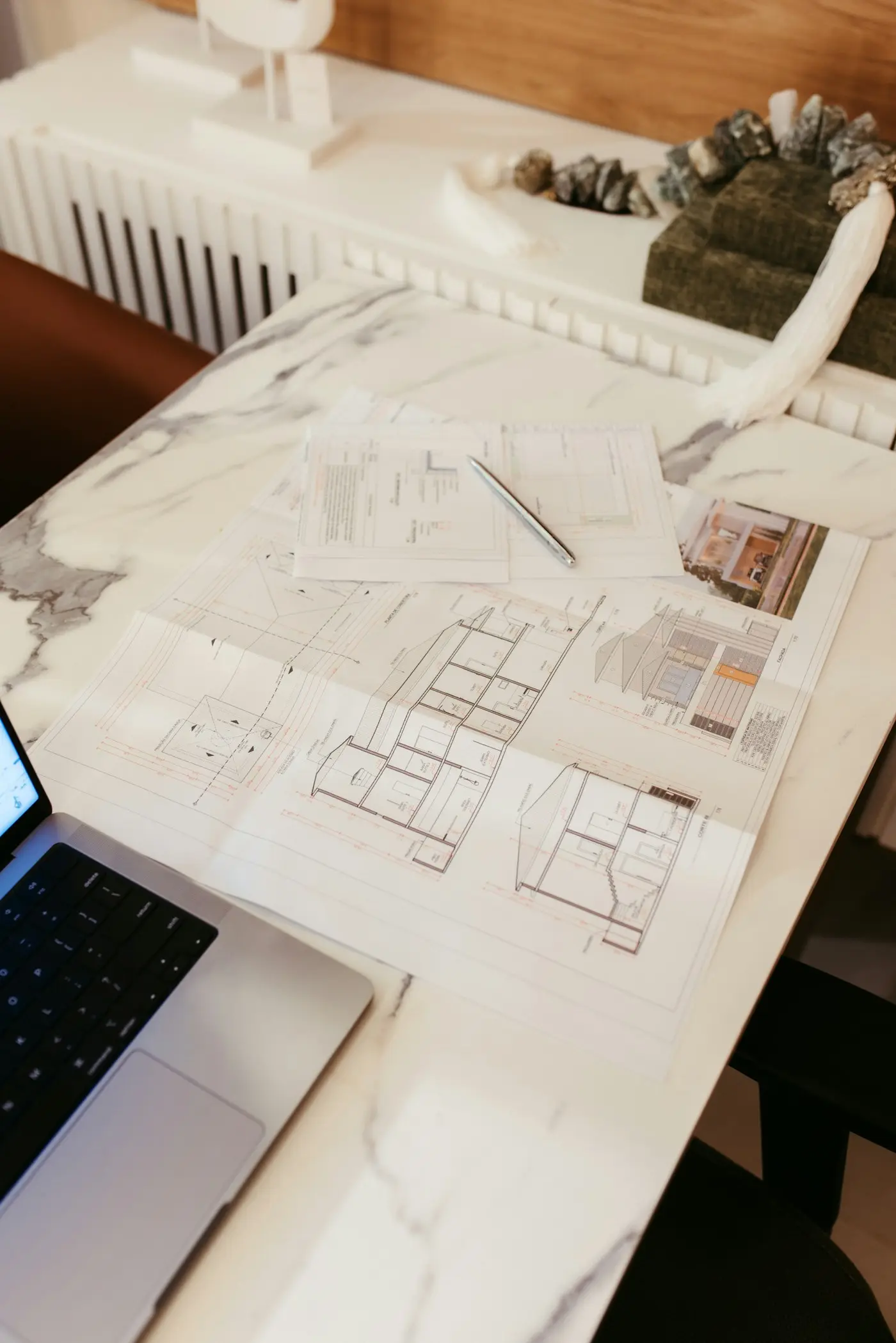 Architectural drawings — sections and a floor plan — spread on a marble desk beside a laptop