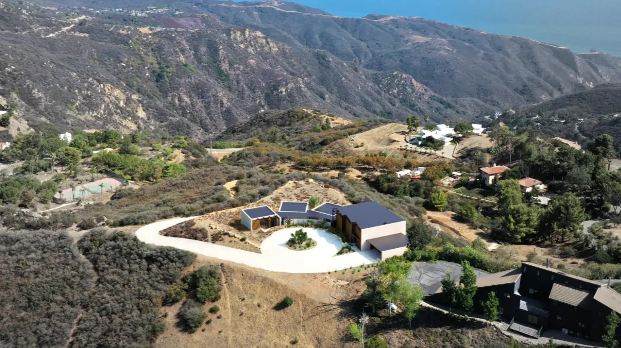 Aerial photograph of the house under construction on a chaparral hillside above the ocean, with a curved concrete driveway